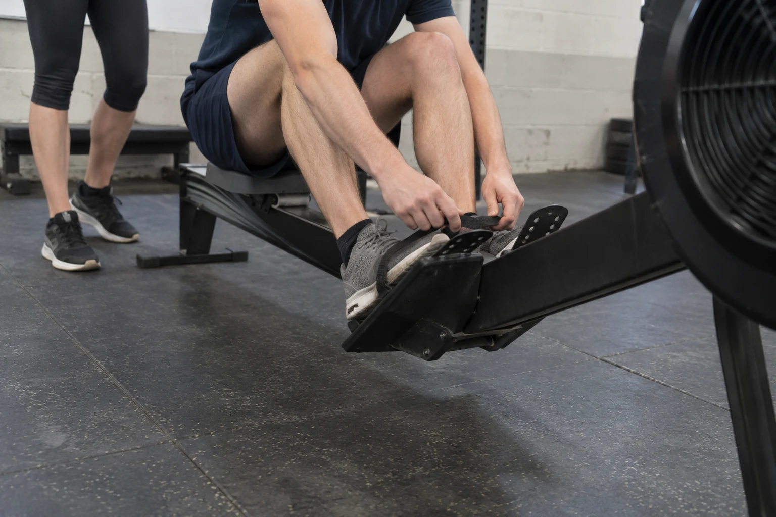 A seated participant adjusts a rowing foot strap while another teammate stands beside the rail